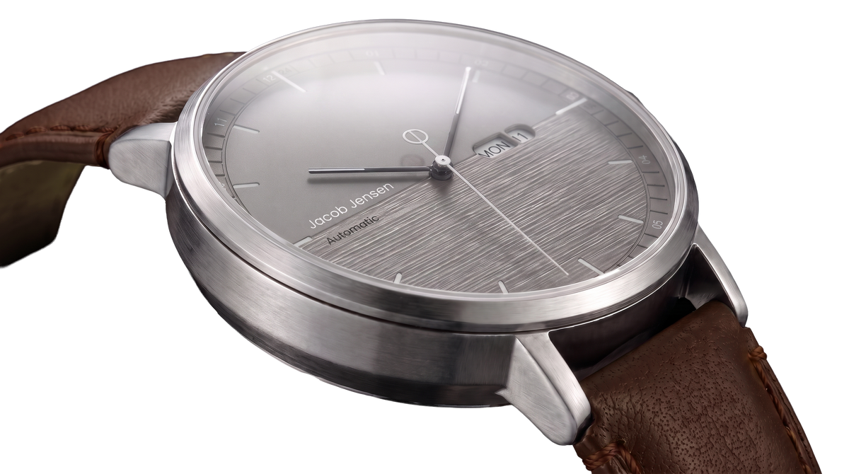A close-up of a Jacob Jensen Automatic watch with a brushed stainless steel case, day-date window, textured gray dial, and a brown leather strap.